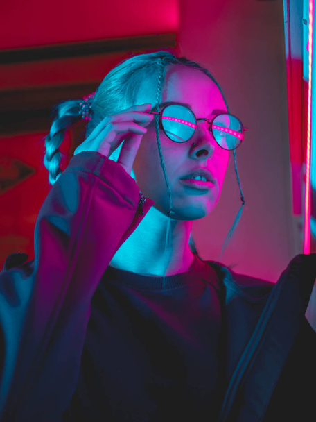 Young pretty girl with unusual hairstyle near glowing pink and blue neon lights of the city at night. Dyed blue hair in braids. Serious hipster teenager in glasses and beautiful lenses. - Fotó, kép