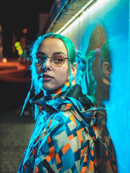 Young pretty girl with unusual hairstyle near glowing turquoise neon light of the city at night. Dyed blue hair in braids. Pensive hipster teenager in glasses. - Foto, afbeelding
