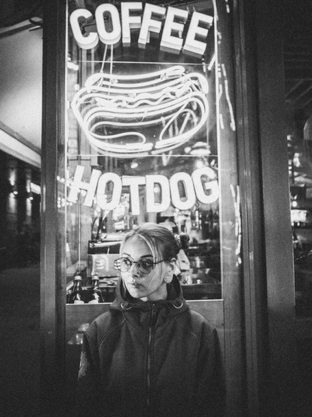 Hipster girl with blue dyed hair. Woman with piercing in nose, lenses, ears tunnels and unusual hairstyle stands at night illuminated city near neon showcase with hot dog - Photo, Image
