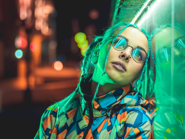 Young pretty girl with unusual hairstyle near glowing green neon light of the city at night. Dyed blue hair in braids. Pensive hipster teenager in glasses. - Foto, Bild