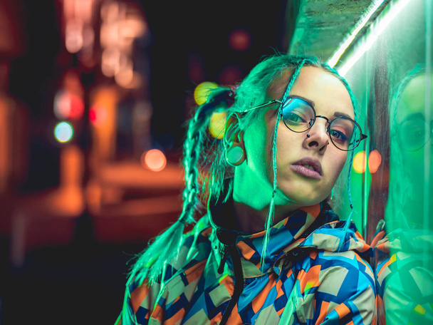 Young pretty girl with unusual hairstyle near glowing green neon light of the city at night. Dyed blue hair in braids. Pensive hipster teenager in glasses. - Photo, image
