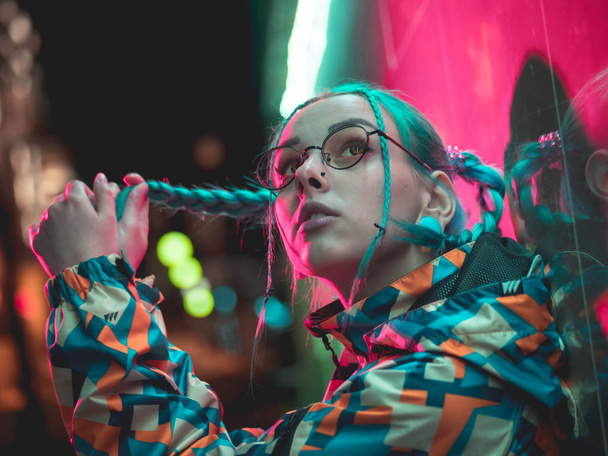 Young pretty girl with unusual hairstyle near glowing crimson red neon light of the city at night. Dyed blue hair in braids. Pensive hipster teenager in glasses. - Foto, afbeelding