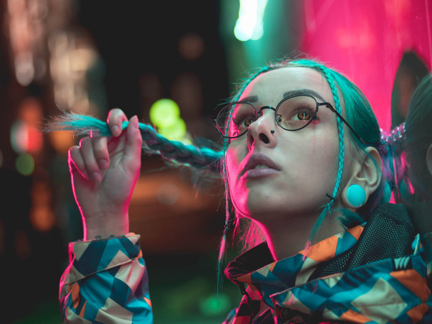 Young pretty girl with unusual hairstyle near glowing crimson red neon light of the city at night. Dyed blue hair in braids. Pensive hipster teenager in glasses. - Foto, Imagem
