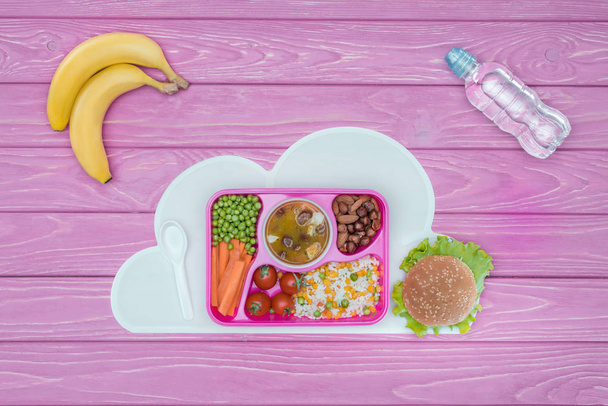 top view of tray with kids lunch for school, bananas and bottle of water on pink table - Фото, изображение