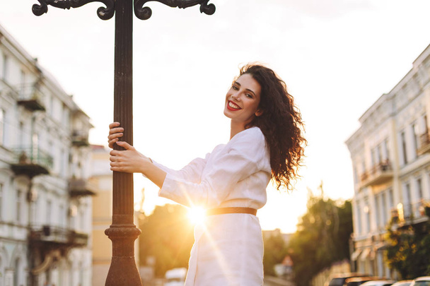Young smiling lady in white jacket standing on street and joyfully looking in camera with beautiful city view on backgroung - Fotoğraf, Görsel