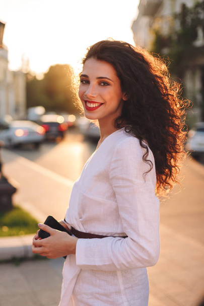 Belle dame souriante en veste blanche debout sur la rue et regardant joyeusement dans la caméra avec téléphone portable dans les mains
 - Photo, image