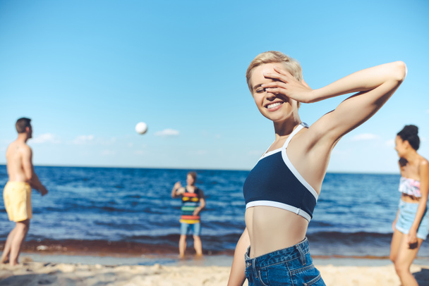 selective focus of smiling woman looking at camera while multicultural friends playing volleyball on beach - 写真・画像