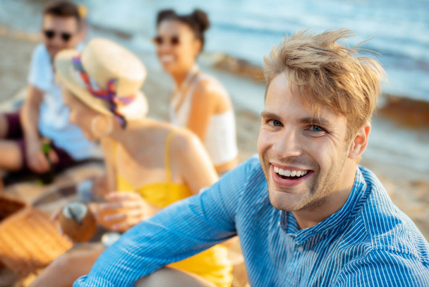 selective focus of cheerful young man looking at camera and multiracial friends near by on beach - Valokuva, kuva