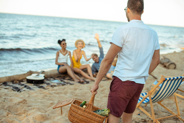 enfoque selectivo del hombre con cesta de picnic y amigos multiétnicos en la playa de arena
 - Foto, Imagen