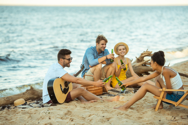 multiethnic smiling friends with drinks and acoustic guitar resting on beach together - Photo, Image