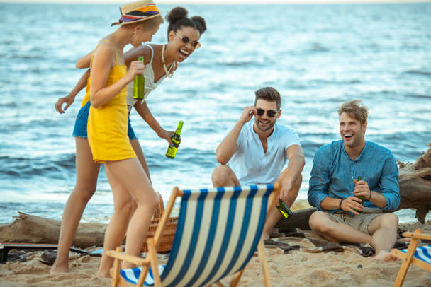 селективное внимание мультикультурных молодых друзей со стеклянными бутылками пива, проводящих время вместе на песчаном пляже
 - Фото, изображение