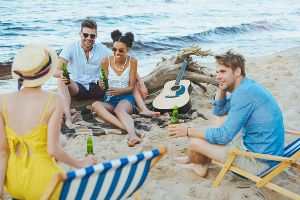 enfoque selectivo de amigos jóvenes multiculturales con botellas de vidrio de cerveza pasar tiempo juntos en la playa de arena
 - Foto, Imagen