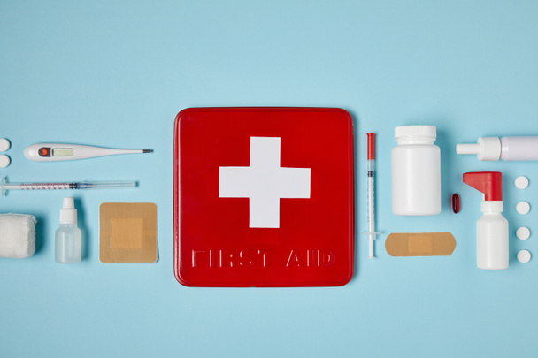 верхний вид красной коробки аптечки на голубой поверхности с медикаментами
 - Фото, изображение