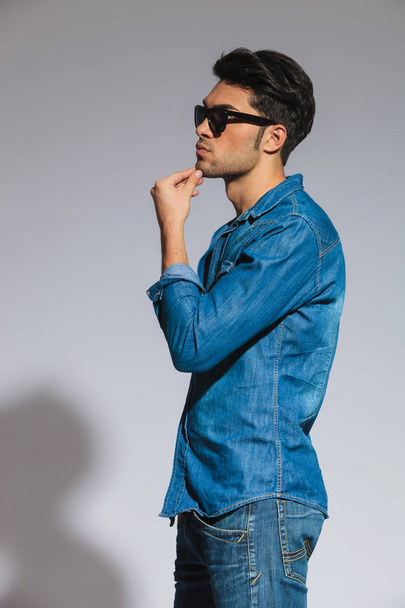 side view of ensive casual man with sunglasses standing on light grey background and looking to side - Photo, image
