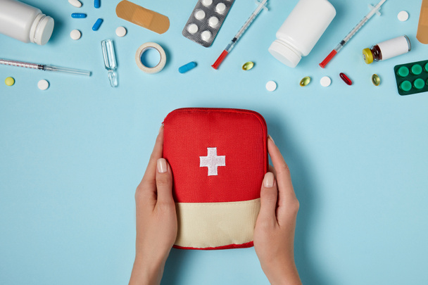 cropped shot of woman holding first aid kit bag over blue surface with various medicines - Photo, Image