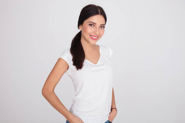 portrait of relaxed brunette woman in white t-shirt standing on light grey background with hands in pockets - Photo, Image