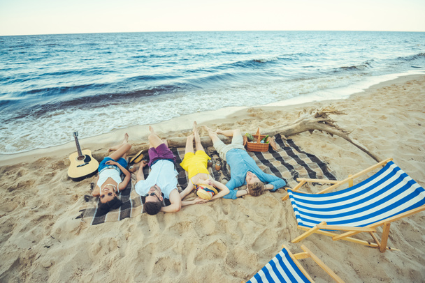 jóvenes multirraciales tumbados en manta mientras pasan tiempo en la playa de arena
 - Foto, Imagen