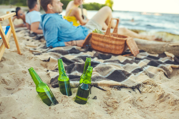 Selektywny fokus szklanych butelek piwa w piasek i Międzyrasowe młodych ludzi odpoczynku w pobliżu przez - Zdjęcie, obraz