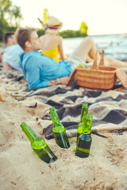 selective focus of glass bottles of beer in sand and interracial young people resting near by - Photo, Image
