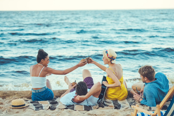 amici multiculturali con cocktail al cocco e chitarra acustica appoggiati sulla spiaggia sabbiosa
 - Foto, immagini