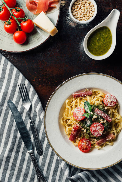 top view of pasta with mint leaves, jamon and cherry tomatoes covered by grated parmesan on plate at table with kitchen towel, knife, fork, pine nuts and pesto sauce in bowl  - Foto, Imagen