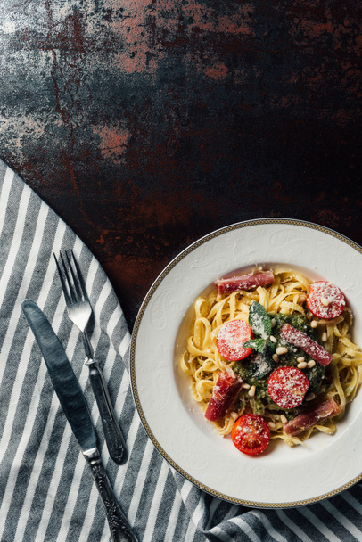elevated view of pasta with mint leaves, jamon and cherry tomatoes covered by grated parmesan on plate at table with kitchen towel, knife and fork - Photo, image
