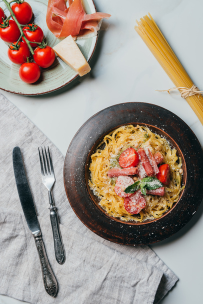 top view of plates with pasta and jamon, parmesan and jamon on marble table with kitchen towel, fork, knife and raw spaghetti  - Fotografie, Obrázek