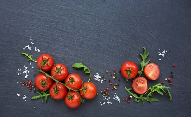 Fresh cherry tomatoes, salt, pepper and arugula leaf on a black slate board, top view - Photo, Image