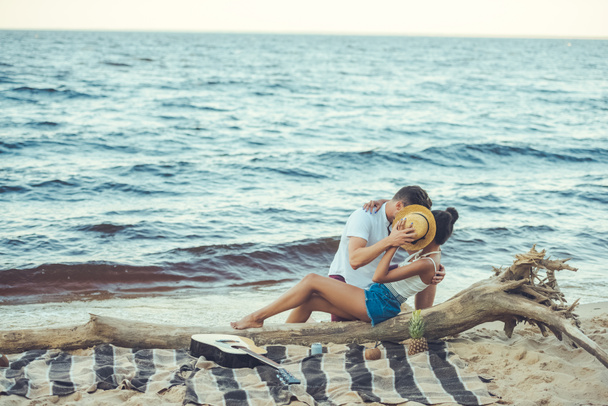 obscured view of multiethnic couple hiding behind hat while kissing on beach - Foto, imagen