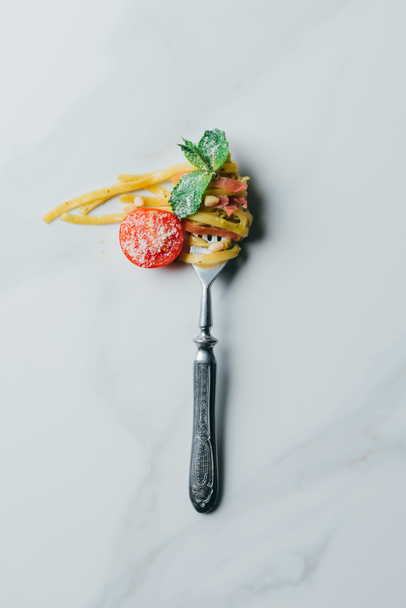 top view of fork wrapped by pasta with mint leaves, jamon and cherry tomatoes covered by grated parmesan on marble table - 写真・画像