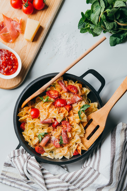 view from above of pasta with jamon, cherry tomatoes, mint leaves covered by grated parmesan in pan with wooden spatula and spoon on marble table - Fotografie, Obrázek