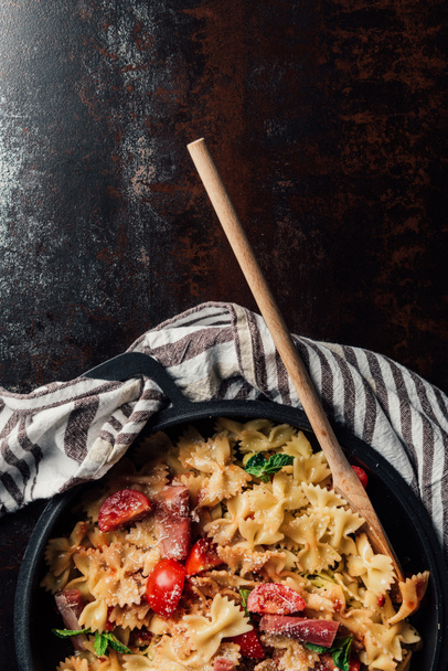elevated view of pasta with jamon, cherry tomatoes, mint leaves covered by grated parmesan in pan with wooden spatula on table with kitchen towel - Foto, Bild