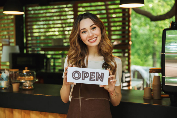 Retrato de una joven barista sonriente en delantal sosteniendo un letrero abierto mientras está de pie en el café
 - Foto, imagen