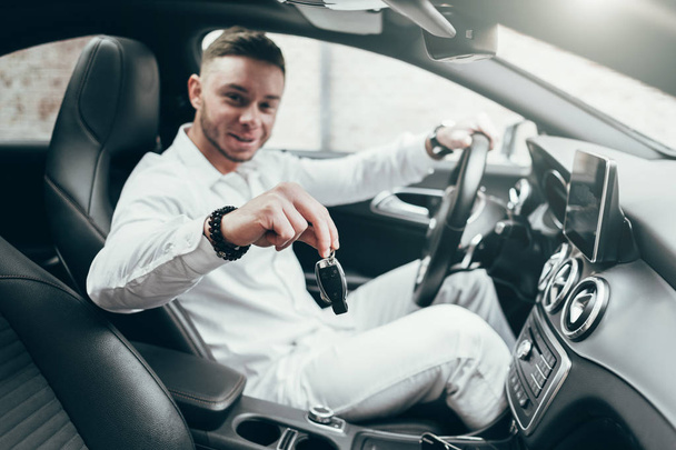 Joven guapo mostrando llaves de coche en su auto recién comprado sentado en el vehículo de lujo
 - Foto, Imagen