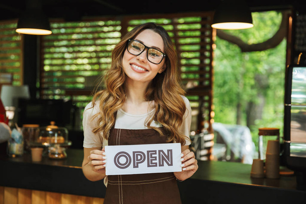 Portrait of a pretty young barista girl in apron holding open sign board while standing at the cafe - Foto, Bild