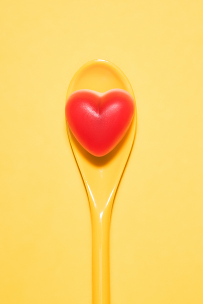 top view of delicious candy in shape of red heart on spoon and on yellow surface - Photo, Image