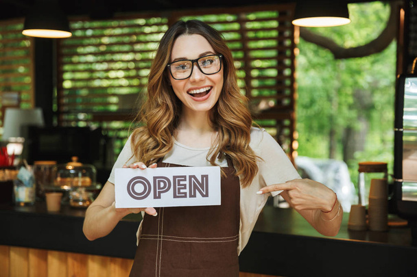Portrait of a smiling young barista girl in apron holding open sign board while standing at the cafe and pointing - Fotografie, Obrázek