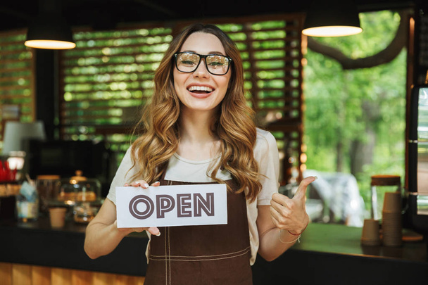 Retrato de una joven camarera sonriente en delantal sosteniendo un letrero abierto mientras está de pie en el café y mostrando los pulgares hacia arriba
 - Foto, imagen