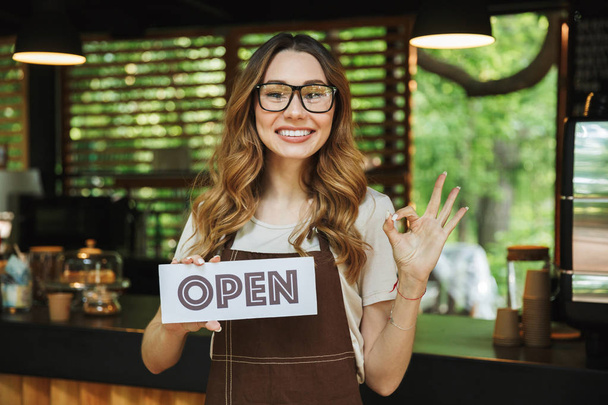 Portrait of a smiling young barista girl in apron holding open sign board while standing at the cafe and showing ok gesture - Foto, immagini