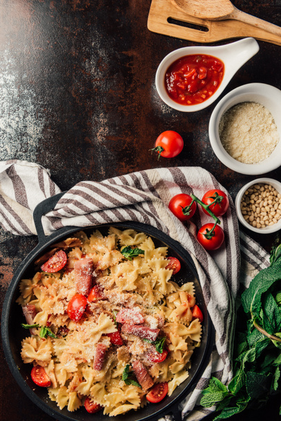 top view of pasta with jamon, pine nuts, sauce, cherry tomatoes, mint leaves covered by grated parmesan in pan surrounded by ingredients and kitchen towel on table  - Foto, Imagen