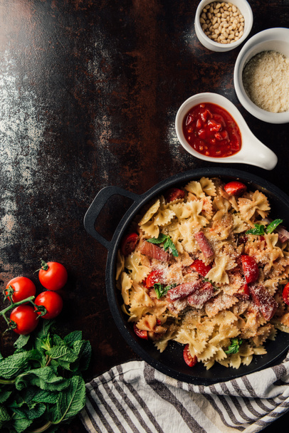 elevated view of pasta with jamon, pine nuts, sauce, cherry tomatoes, mint leaves covered by grated parmesan in pan surrounded by ingredients and kitchen towel on table  - Foto, Imagen