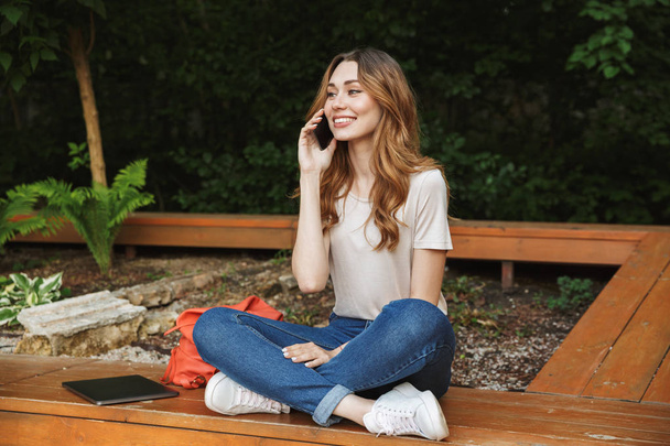 Smiling young girl talking on mobile phone while sitting on a bench outdoors - Foto, Imagen