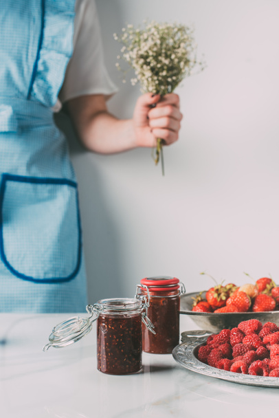 cropped image of woman in apron holding flowers near table with raspberries, strawberries and jars of jam  - Photo, image