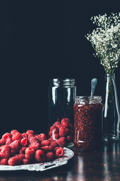 close up view of raspberries in silver tray, flowers, jar with jam on black  - Photo, Image