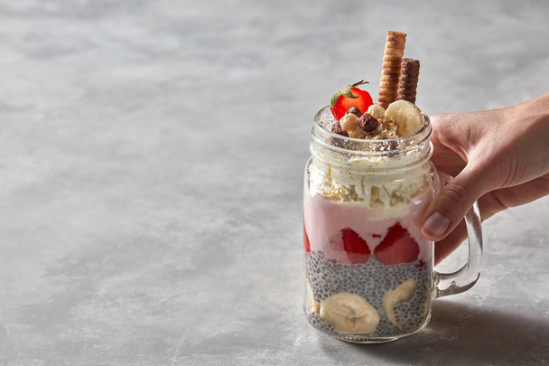 A glass with a handle in a woman's hand with a healthy breakfast of yogurt, strawberry, chia pudding with a banana, decorated with biscuits and whipped cream on a gray concrete background. Copy space - Фото, изображение