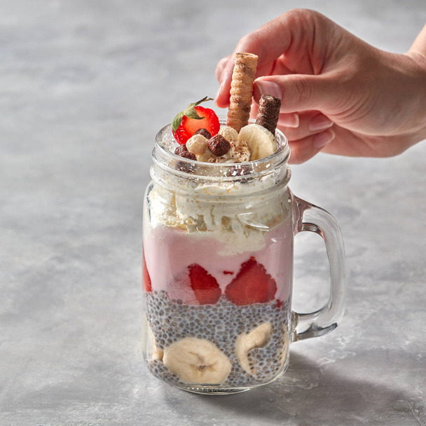 Woman's hand decorates healthy layered dessert with yogurt, chia pudding, banana and strawberry in glass on a stone background - 写真・画像