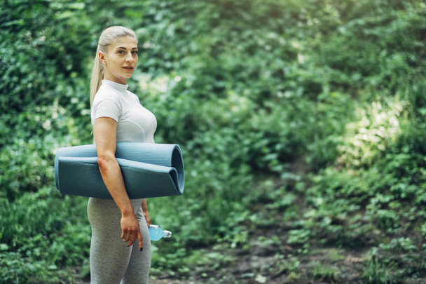 Женщина готовится к Йоге в парке ранним утром. Носит стильный спортивный костюм. Голубая бутылка воды и коврик для йоги. Здоровый образ жизни
. - Фото, изображение