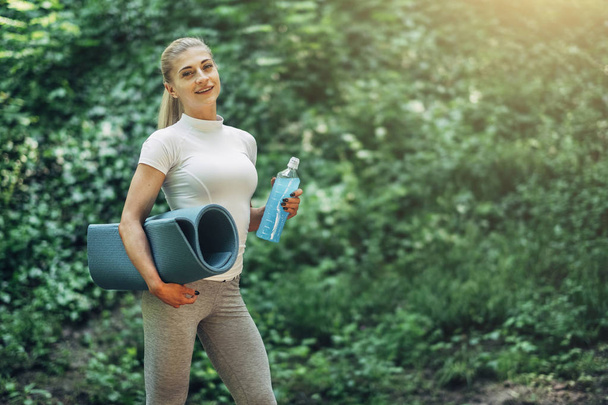 Mujer Fitness Preparándose para el Yoga en el Parque Temprano en la Mañana. Usando ropa deportiva con estilo. Botella Azul de Agua y Estera de Yoga. Estilo de vida saludable
. - Foto, Imagen