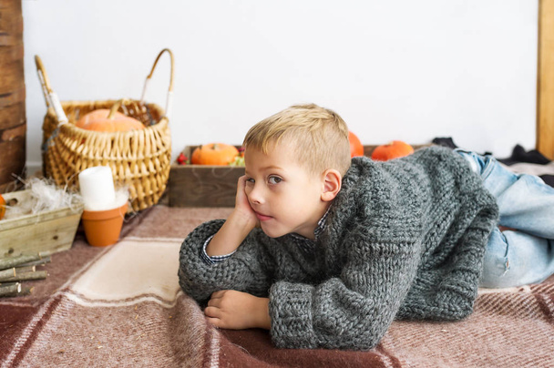 Portrait of the quiet lovely boy lying on a cover in the home room against the background of orange pumpkins - Foto, Imagen