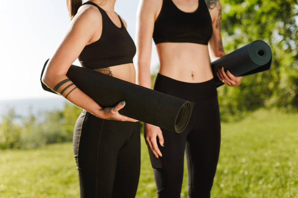Close up photo of women bodies in black sporty tops and leggings standing and holding yoga mats in hands with beautiful view on background - 写真・画像